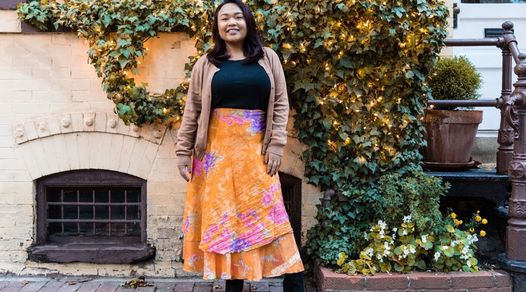A woman in an orange skirt with purple accents, standing in front of walls and fairy lights