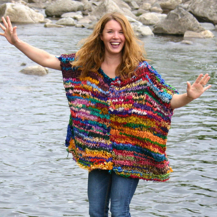 A woman with a big smile standing on the river wearing a colorful poncho knit shirt.