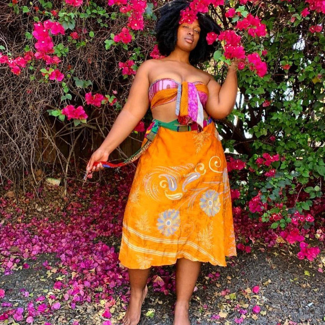Goddess Model wearing an orange tea length sari wrap skirt in front of bright pink flower bush.