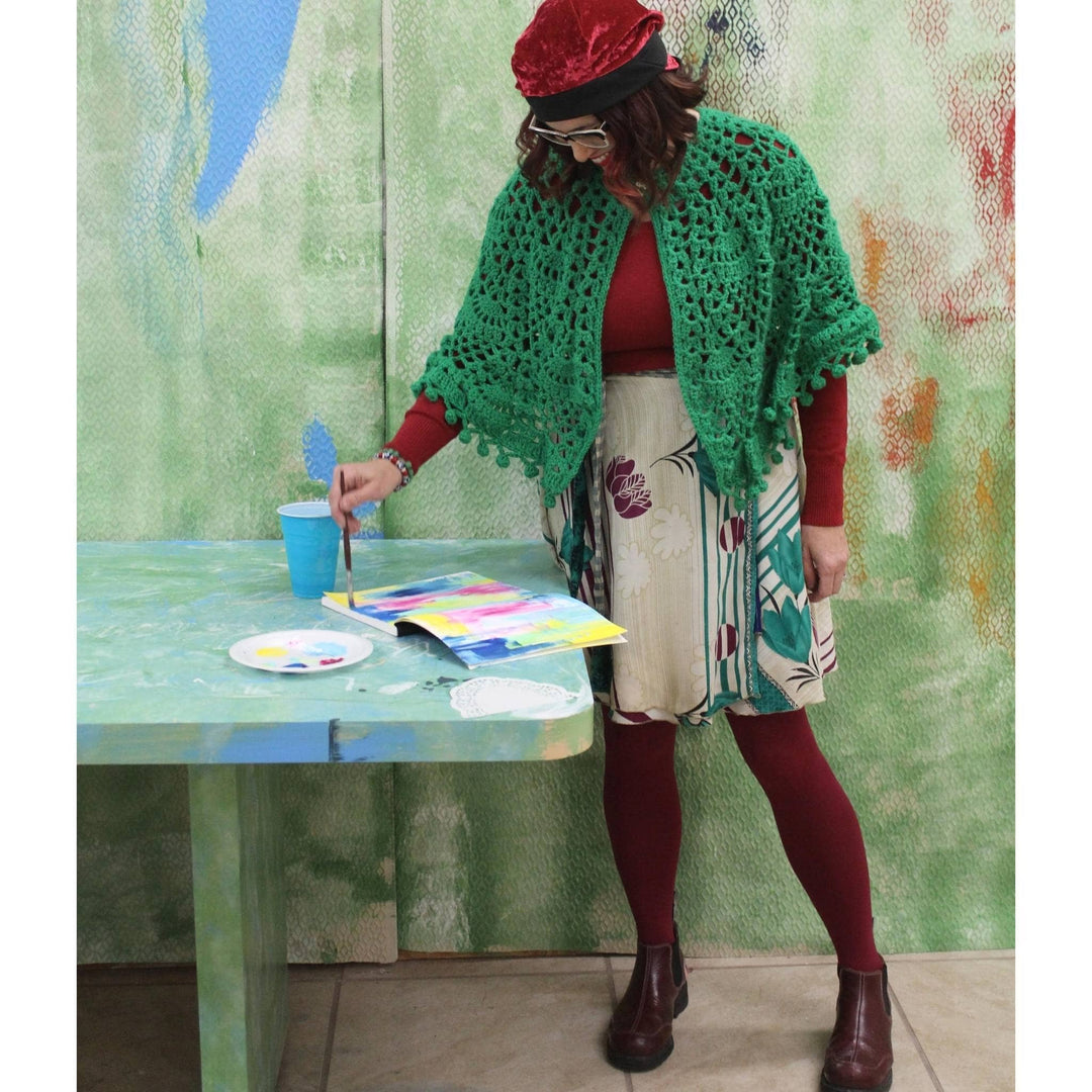 Artist Deanna Bowers wearing a white and turquoise mini sari wrap skirt. She is standing in front of a green wall, painting at a green table.