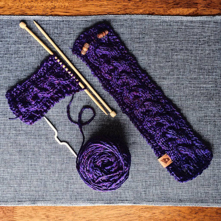 Purple knitted cozy with wooden buttons with purple yarn cake and knitting needles sitting on a gray fabric surface