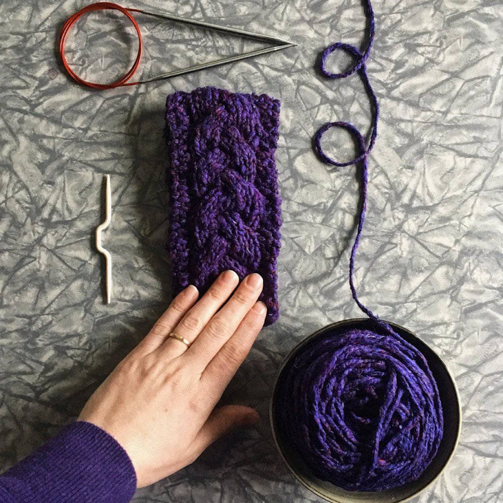 Purple knitted cozy with wooden buttons with purple yarn cake, circular knitting needles, and womans hand on a gray surface