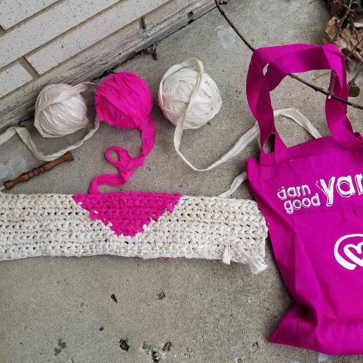 Partially made All My Love crochet Wall Hanging sitting next two 3 balls of cream and pink yarn and a hot pink Darn Good Yarn tote bag on a concrete surface