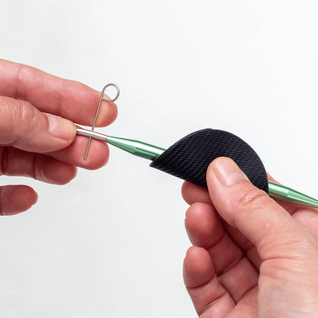 Image of interchangeable knitting needle being assembled in front of a white background. Hand using grip on green aluminum knitting needle, while other hand uses turn key took to tighten cord. 