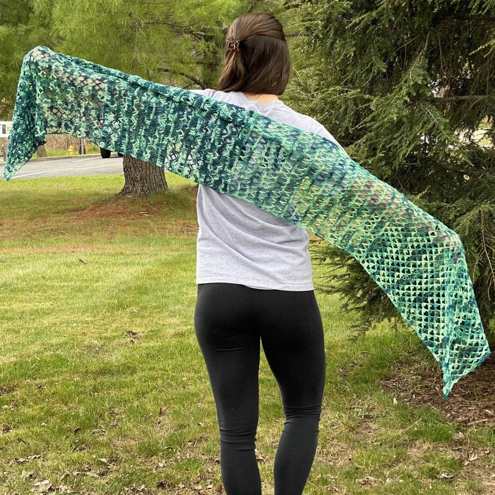 model holding out ariel shawl in variegated green colorway with greenery in the background.