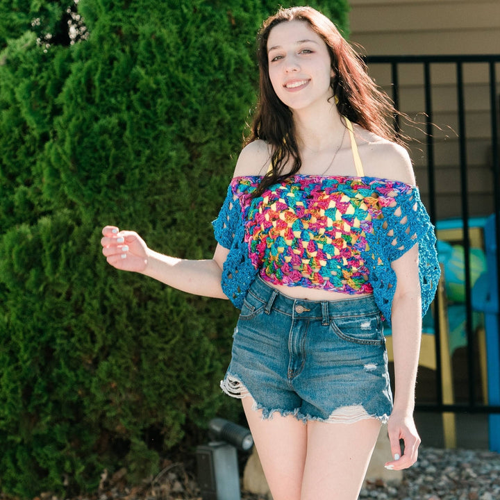 Model wearing blue and multicolor crochet granny square top with greenery in the background.