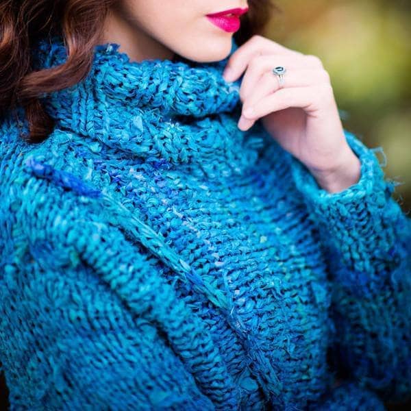 Closeup of woman wearing the blue Catwalk Sweater with cowl neck and crossed front standing by greenery