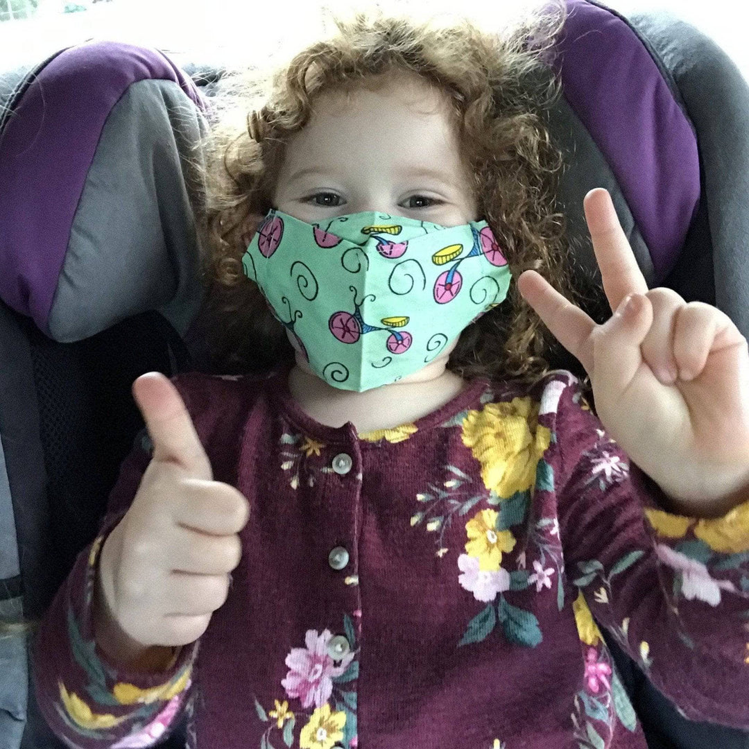 approx 4 year old child wearing green face mask with hands forming a thumbs up and peace sign. 