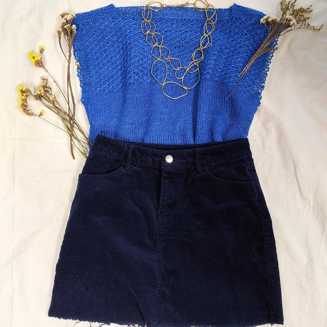 daisy stitch short sleeve knit top in colorway cadet blue, black denim skirt, and a gold necklace laying on a white background with dried flowers around.
