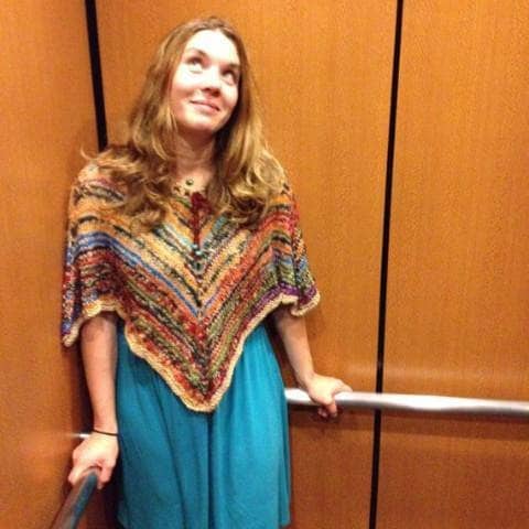 Woman standing in the corner of a brown elevator and wearing a blue dress with multicolored knit poncho