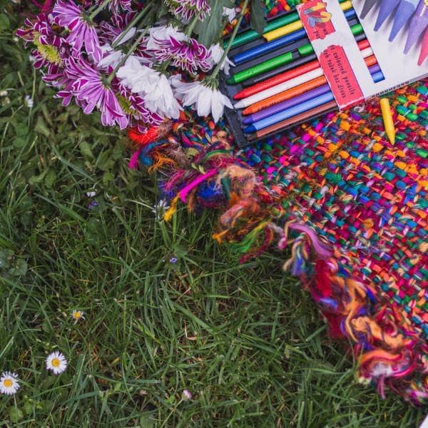 Multicolored woven blanket topped with fresh flowers and colored pencils all laying on the grass