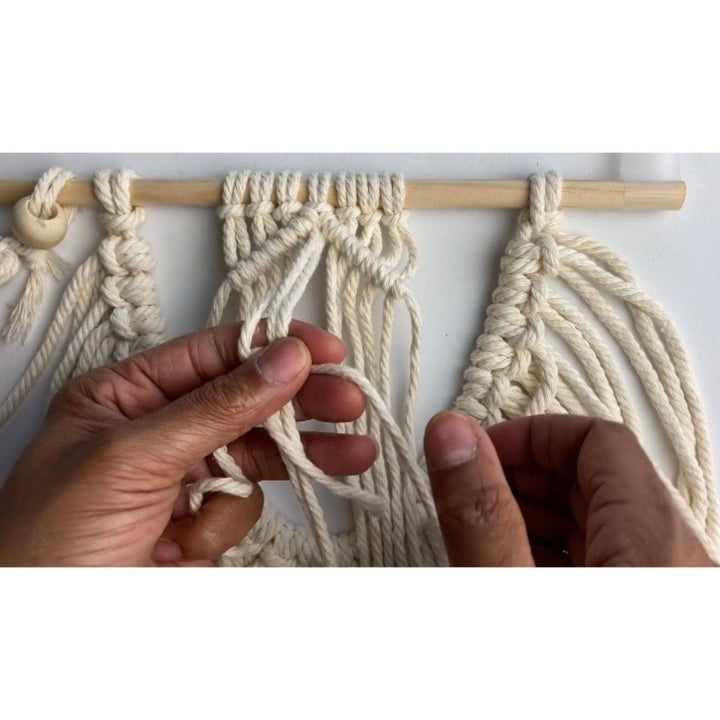 A macrame wall hanging being assembled by skilled hands in front of a white background. 