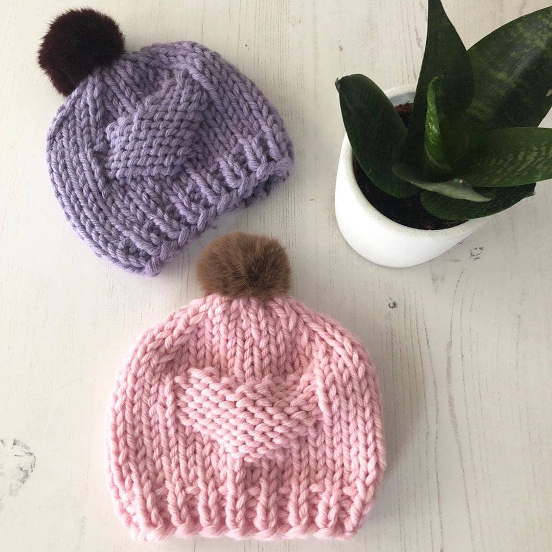 One purple and one pink kids pom pom knit hats on a wooden background. A potted plant is to the right of them. 