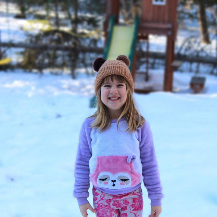 Small child in pink and purple outfit wearing brown knit hat with two pom poms on top standing in front of a snowy playset outside.