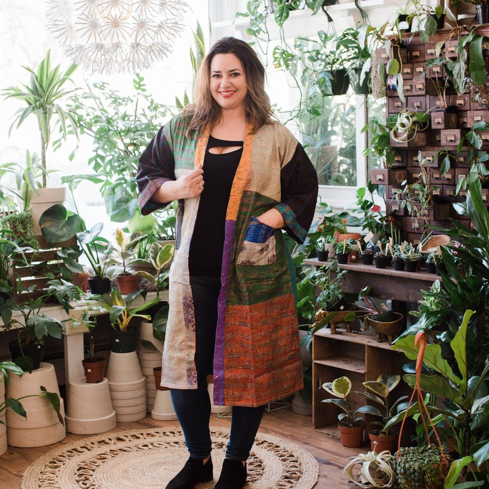 Model is wearing a multicolored kantha jacket while standing in front of some potted plants.   