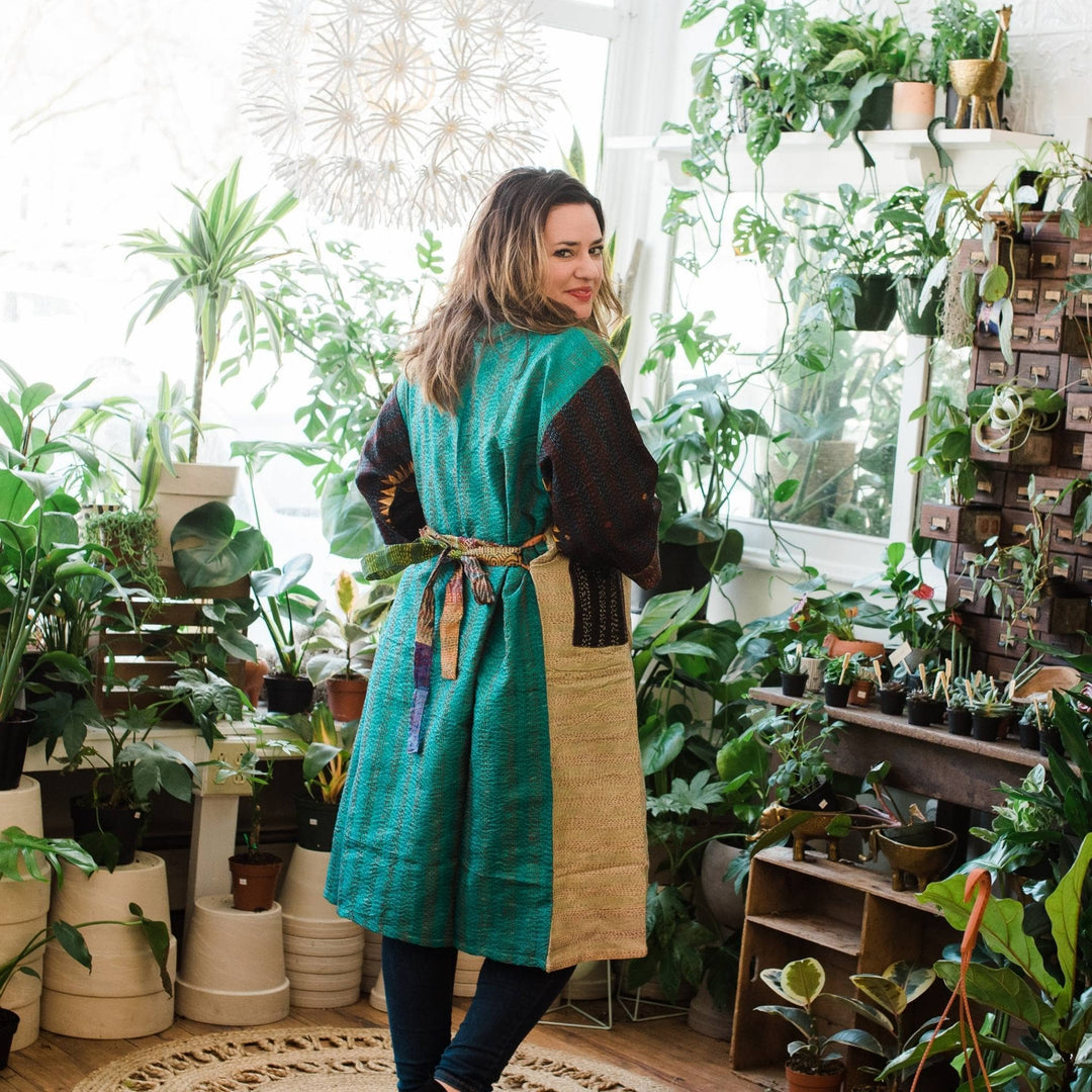 Back of model wearing a teal kantha jacket tie in the back. Potted plants are in the background. 