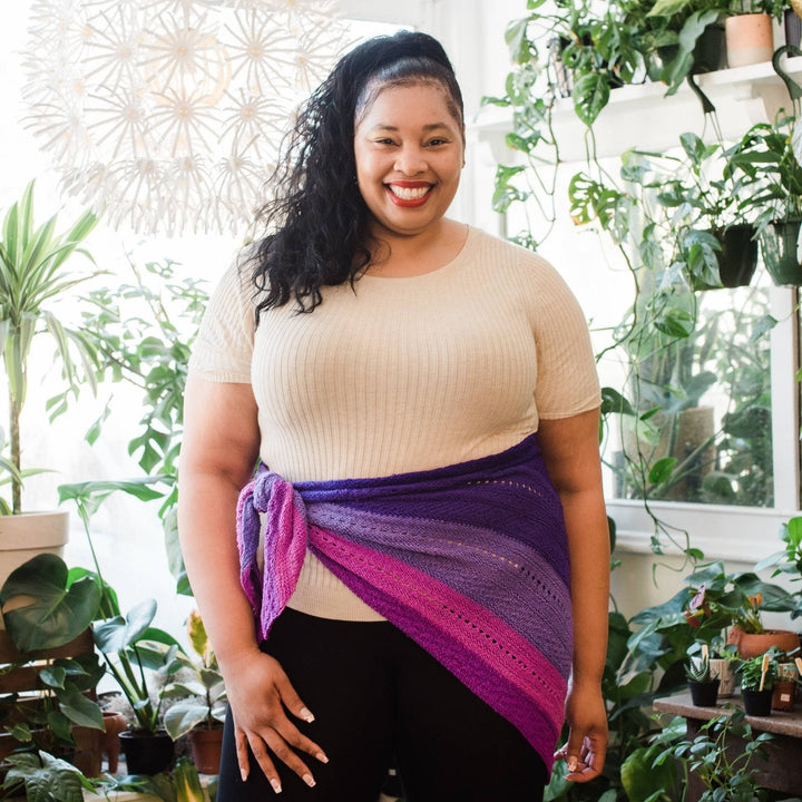 Model wearing ombre stitch sampler shawl tied around the waist with potted greenery in the background.