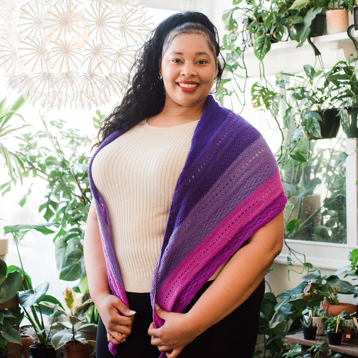 model wearing purple ombre stitch sampler shawl over their shoulders with potted greenery in the background.