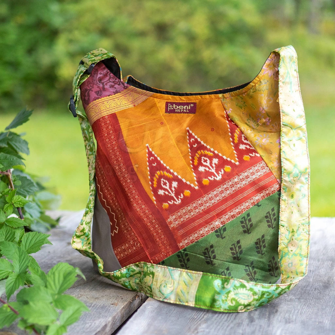 One of a kind sari purse sitting on a wood surface next to a leafy bramble with greenery in the background. Purse is multicolor with a red tag that reads beni. 