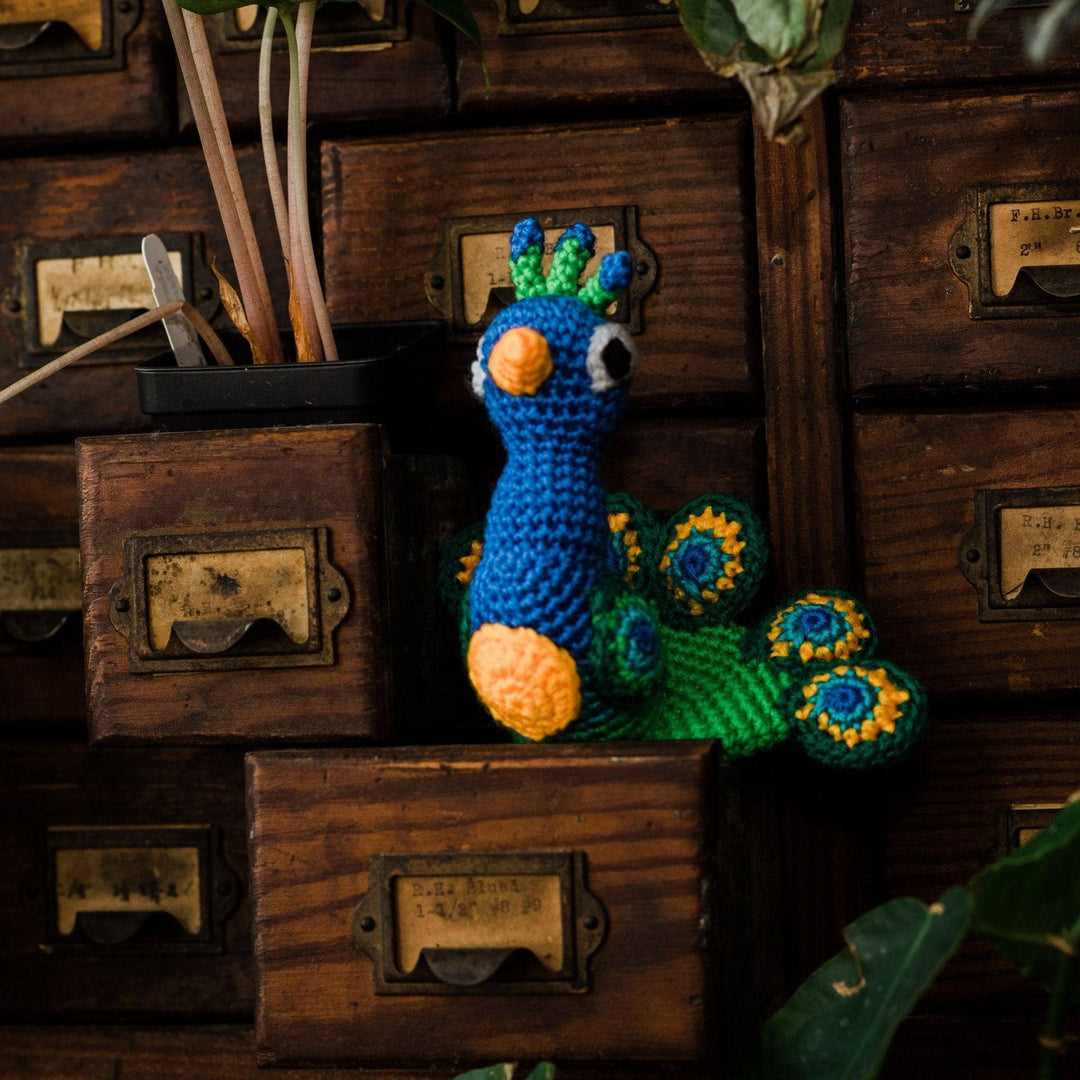 Completed peacock amigurumi sitting on open apothecary drawer beside other apothecary drawers and potted plants. 
