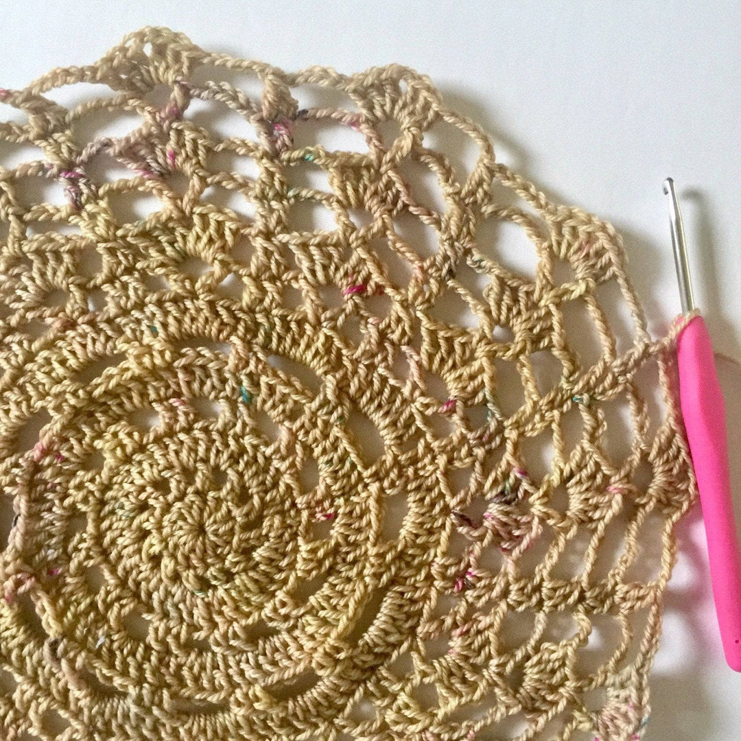 close up of a crochet doilie over a table with a pink hook next to it