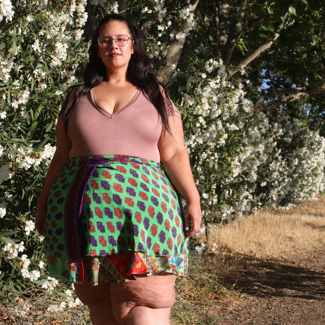 Model is outside in front of a white flower bush wearing a goddess mini sari wrap skirt in green. 