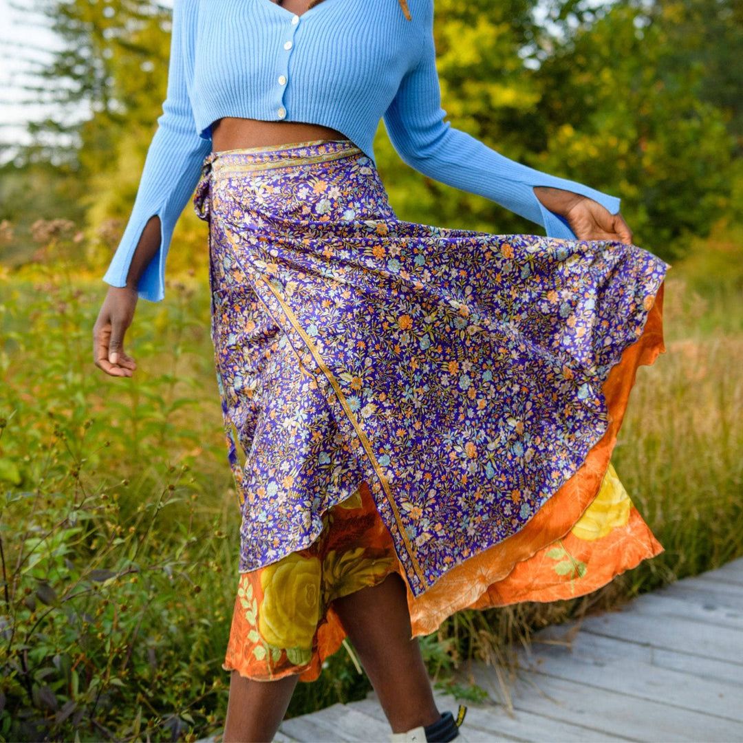 An up close shot of a woman on a board walk wearing a purple floral sari wrap skirt. The under layer is bright orange with gold flowers. She's paired it with a baby blue cropped sweater.
