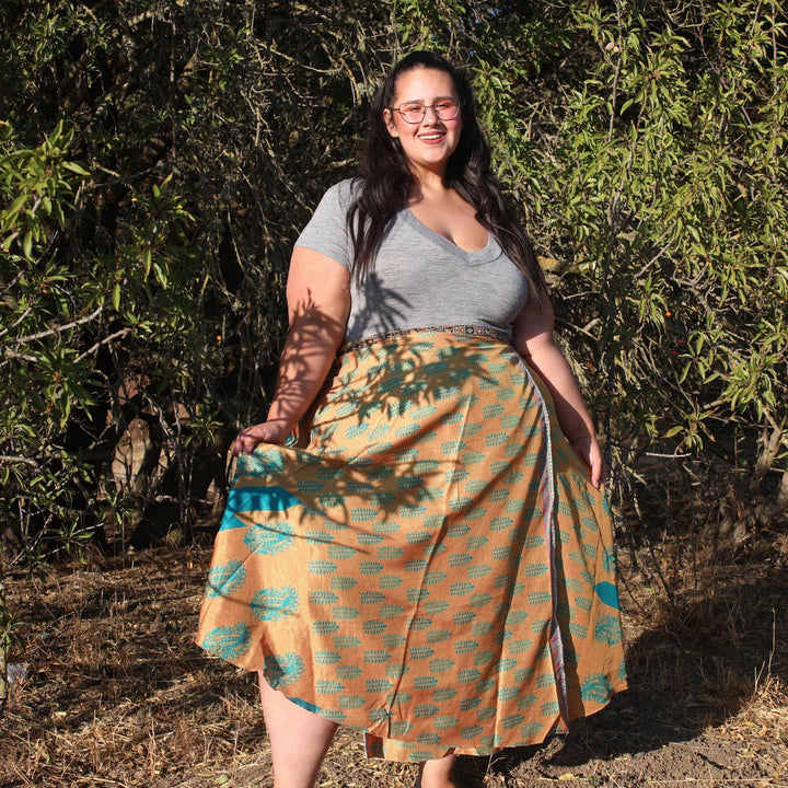 Goddess Model wearing a light orange and blue sari wrap skirt while standing in front of a tree. 
