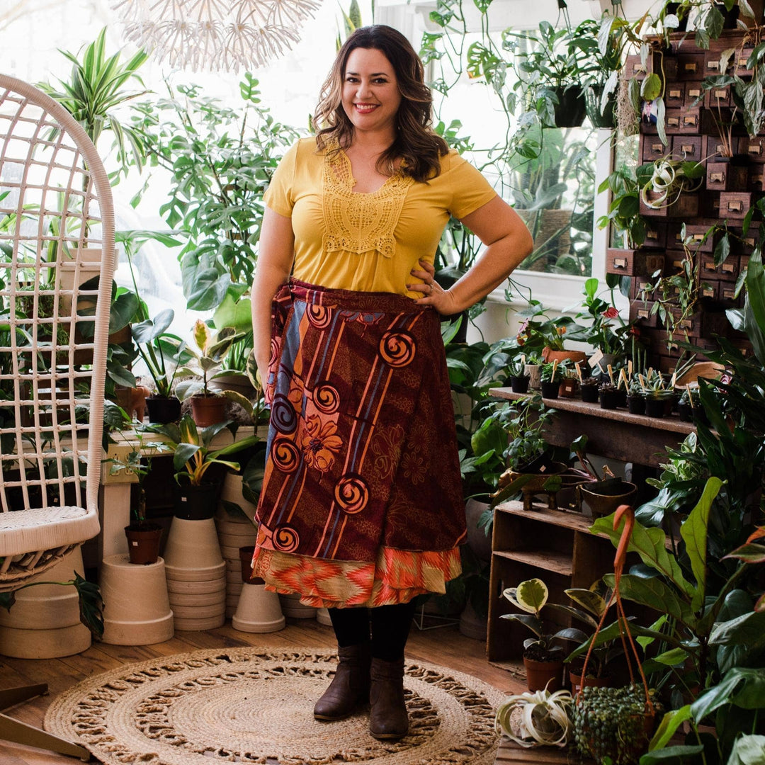 Model is wearing a maroon and orange sari wrap skirt with potted plants in the background.  