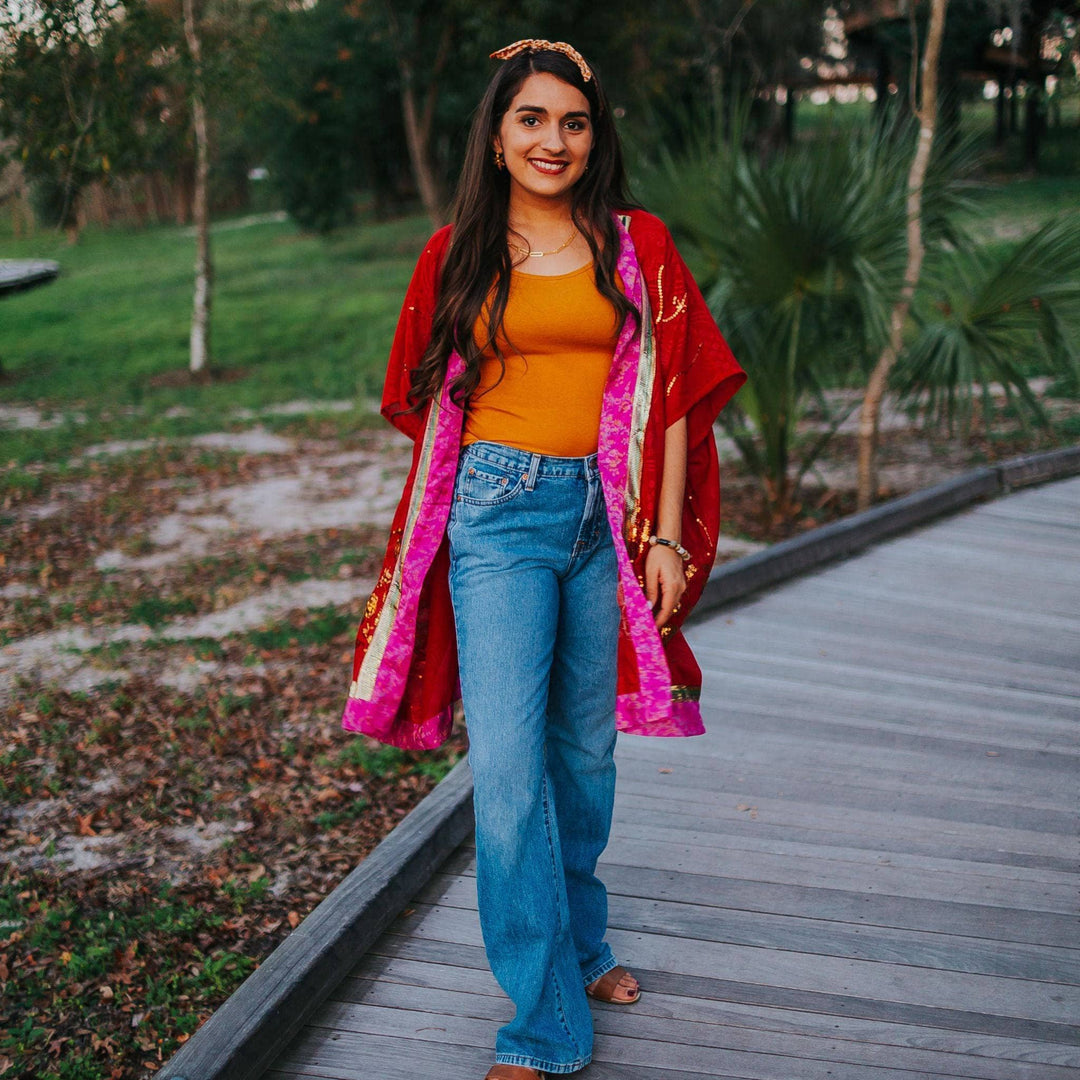 A model standing on a boardwalk at dusk wearing a rich red sophia silk duster with a pair of jeans and a tea. Her duster has vibrant pink edges and beaded floral designs all over. She's paired this look with a kameela knot headband and tan sandals.