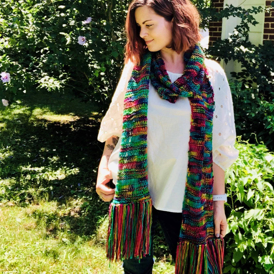 designer wearing iris knit scarf in the colorway watercolors with white top and black pants standing outside with greenery in the background.