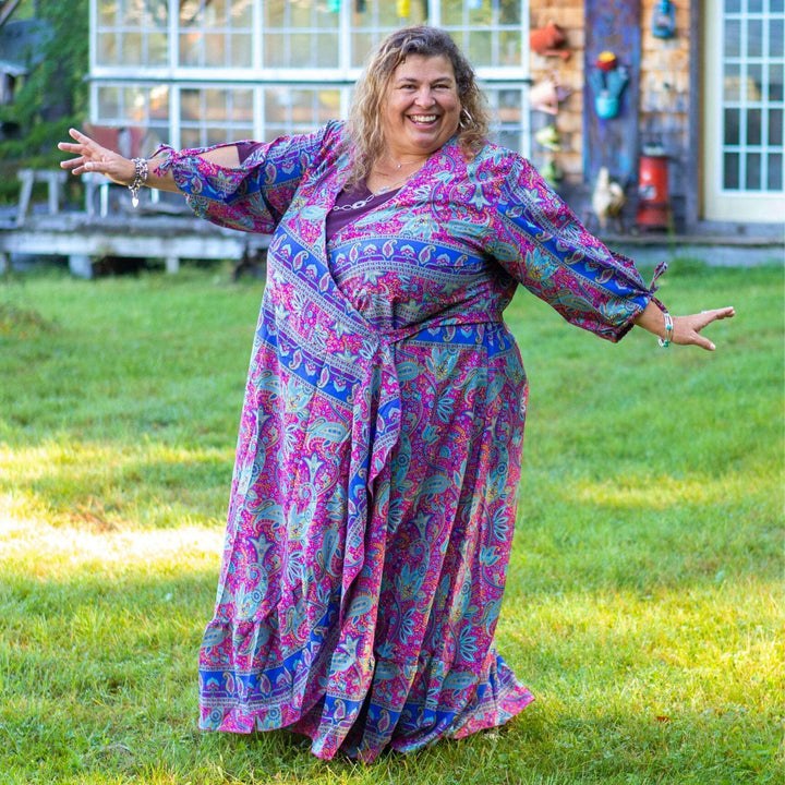 A woman standing in her yard wearing a 14-32 sized zaria wrap dress. Her dress is a purple and light blue paisley whit navy stripes.