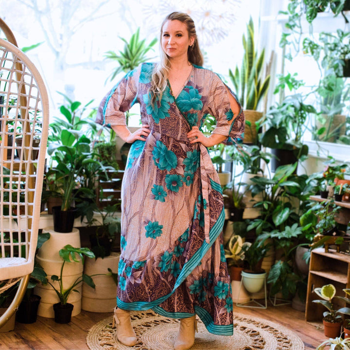 A model in a flower shop wearing a zaria sari wrap dress and cream colored bootie. The dress is a gray base with light blue roses all across it. 