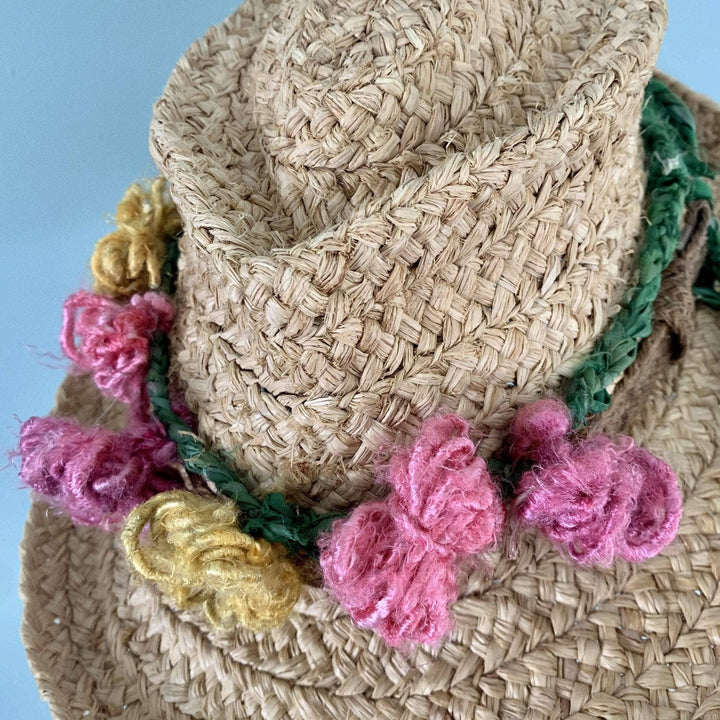 Yarn flower crown worn on a straw hat in front of a light blue background.