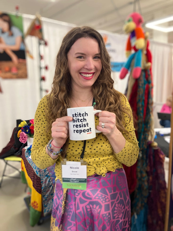 A woman is smiling and holding a mug that says "stitch bitch resist repeat." She is wearing a yellow polka dot top and a colorful skirt. Her name tag reads "Nicole Snow." The background shows a booth with colorful textiles and decorations.