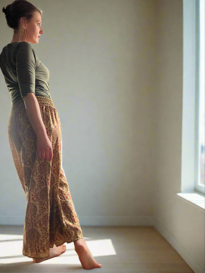 A woman stands barefoot on a wooden floor, leaning against a door. She is wearing a green top and patterned wide-leg pants. Sunlight streams through a window with colorful curtains, casting light on her. There are two potted plants on the floor near the window.