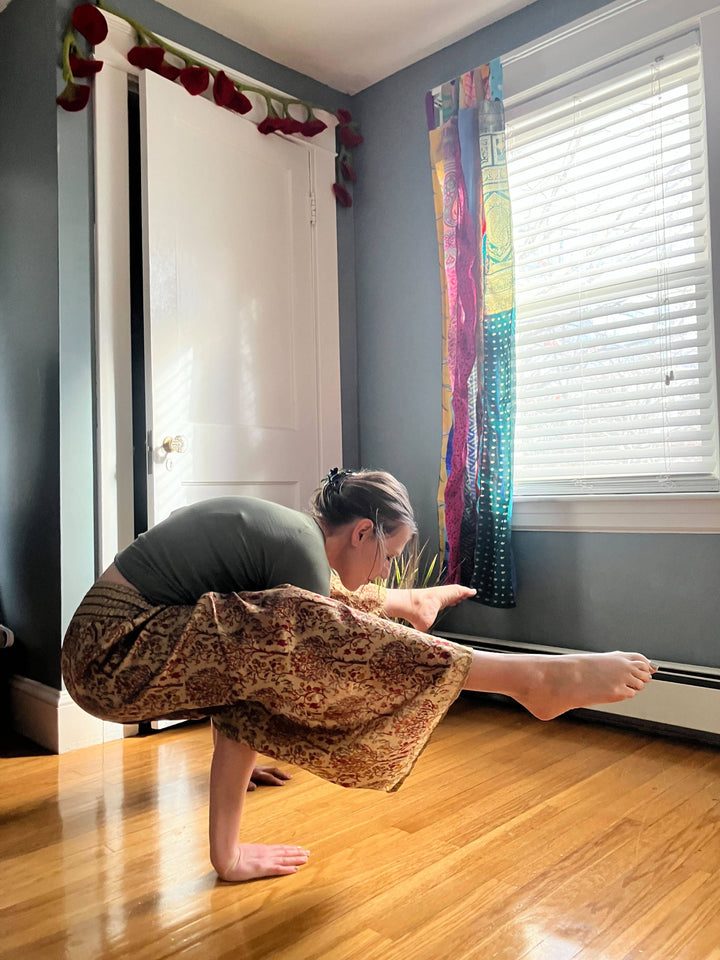 A person is performing a yoga pose indoors, balancing on their hands with one leg extended forward and the other bent. They are wearing a green top and patterned pants. The room has a wooden floor, a white door with red decorations above it, and colorful curtains by a window.