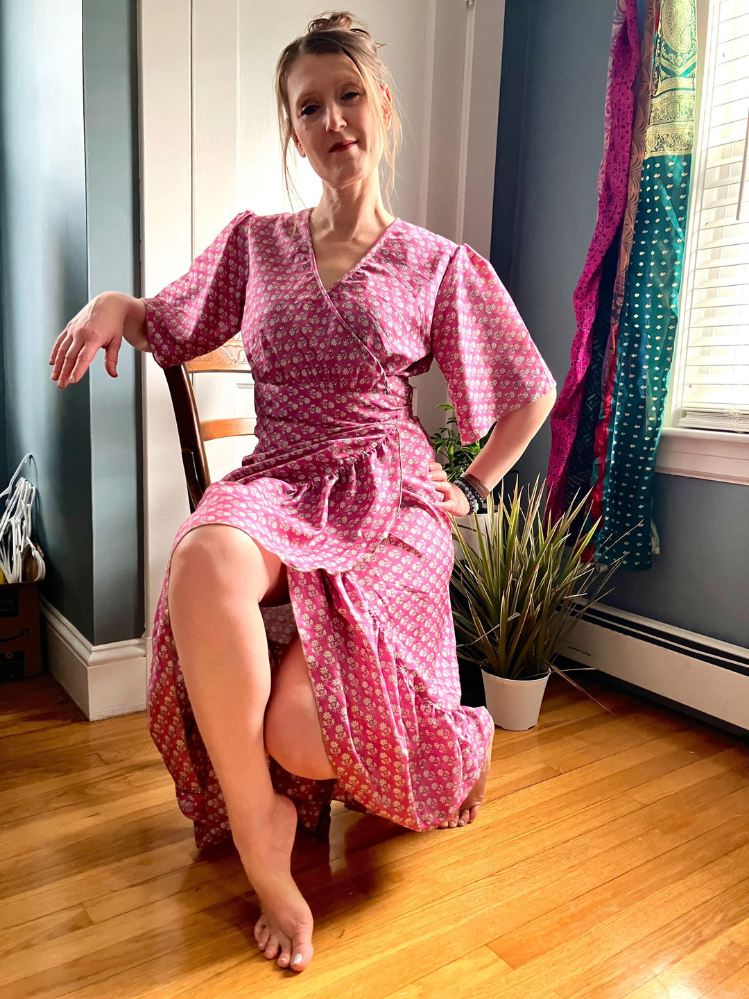 A person sitting on a chair wearing a pink floral wrap dress. The room has wooden floors, a potted plant, and colorful curtains by the window. The person has a relaxed pose with one leg crossed over the other.