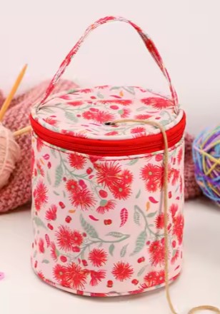 A floral-patterned cylindrical yarn bag with a red zipper and handle. The bag is surrounded by knitting supplies, including yarn balls and knitting needles.