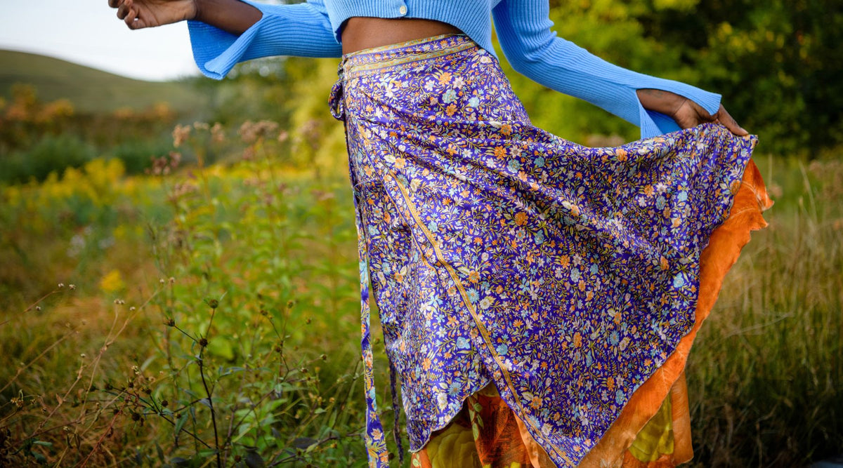 Woman standing in field of green grass with dark purple and orange sari wrap skirt on around dusk. 