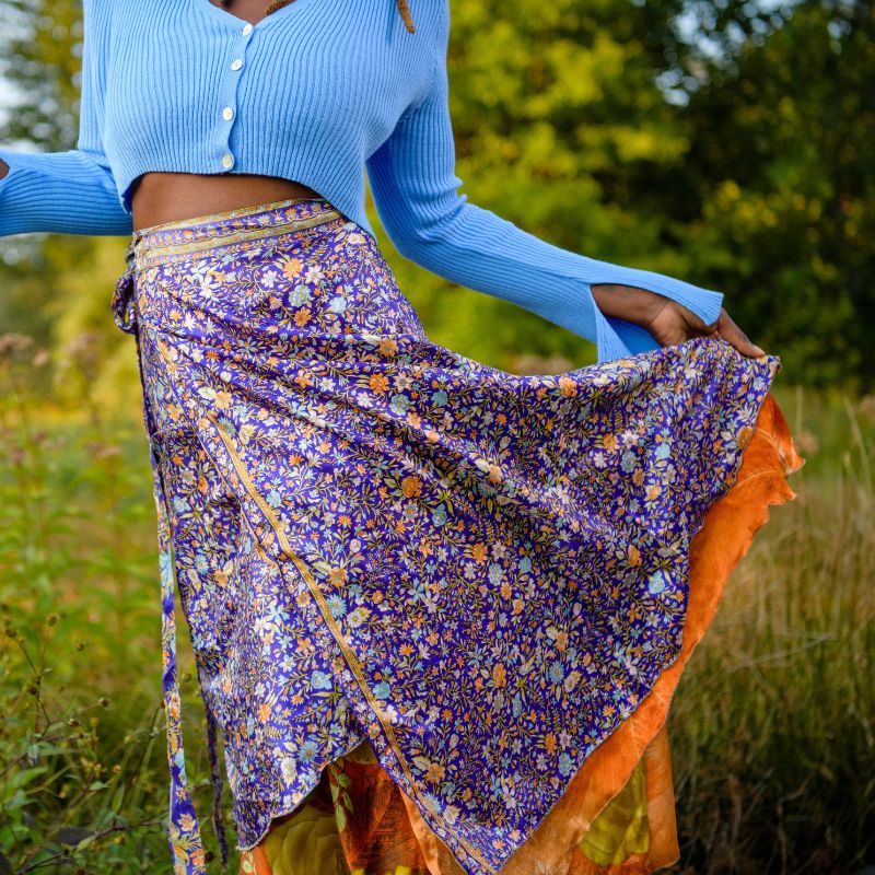 Woman standing in field of green grass with dark purple and orange sari wrap skirt on around dusk. 