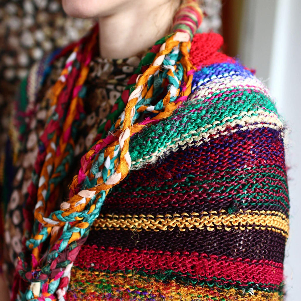 A close-up of a person wearing a colorful, knitted shawl. The shawl features a variety of vibrant colors, including red, green, yellow, and blue, with a braided multicolored pattern along the edge. The background is blurred, focusing attention on the intricate textures and patterns of the shawl.