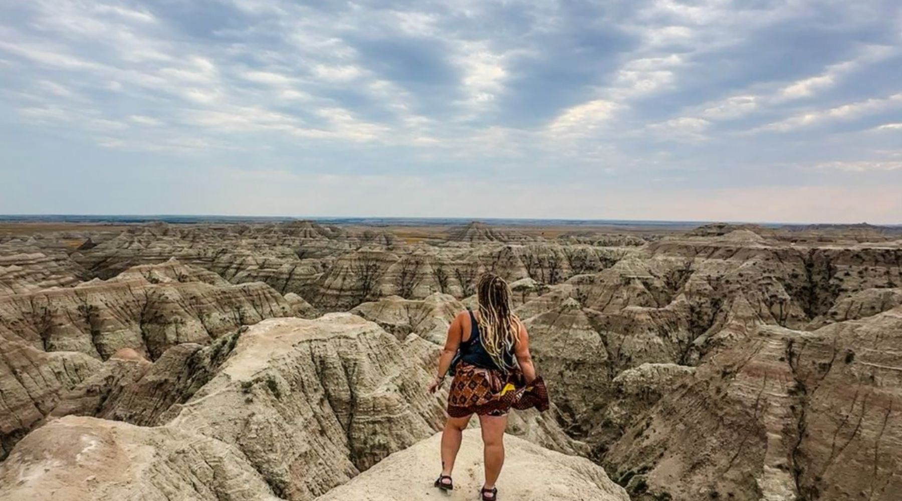 A person with long braided hair stands on a rocky cliff, overlooking a vast landscape of rugged, eroded hills and valleys under a cloudy sky.