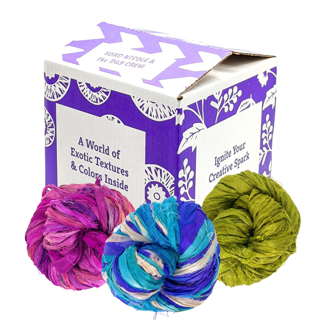 A purple Darn Good Yarn branded box sitting on a white background with three cakes of reclaimed sari silk ribbon yarn sitting in front of it.
