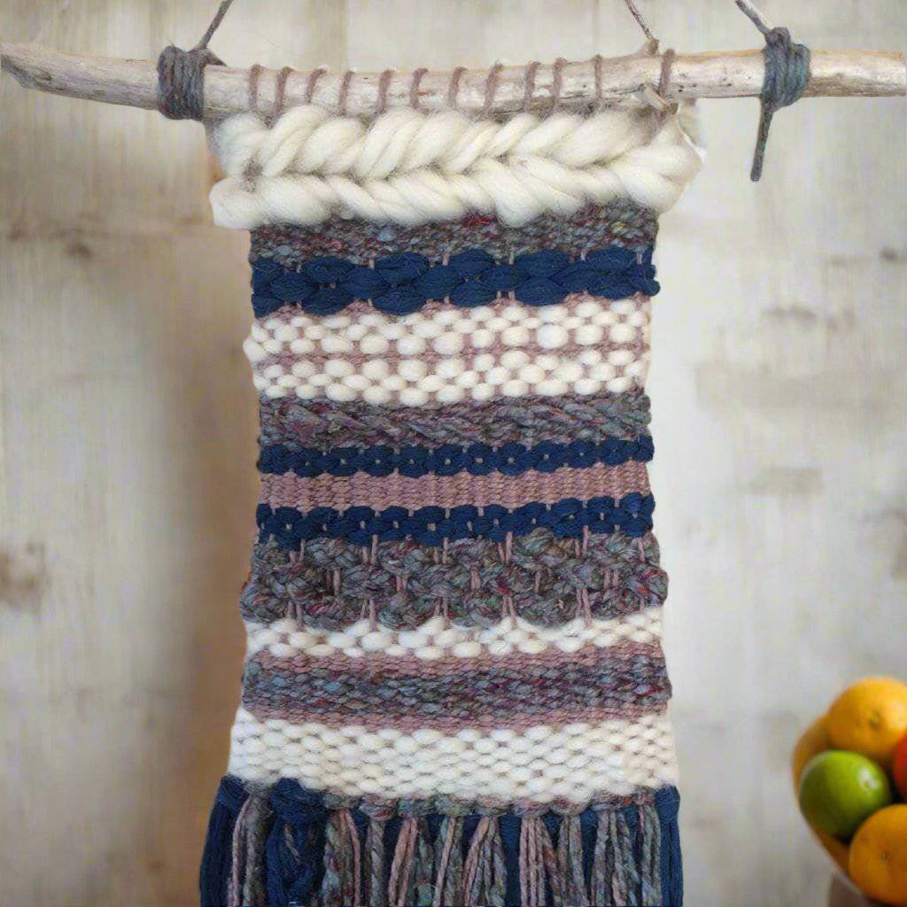 Woven tapestry in gray, navy, and white hanging from wooden stick on a white wall