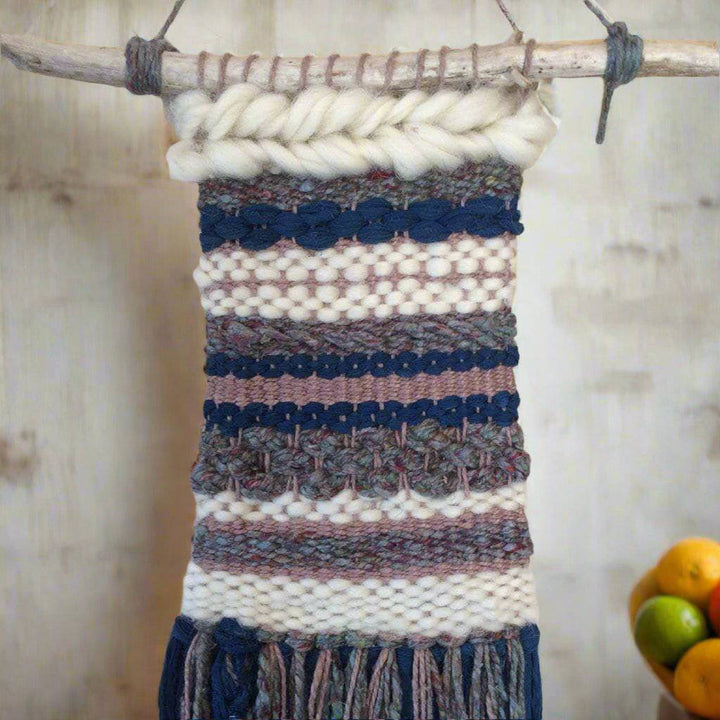 Woven tapestry in gray, navy, and white hanging from wooden stick on a white wall