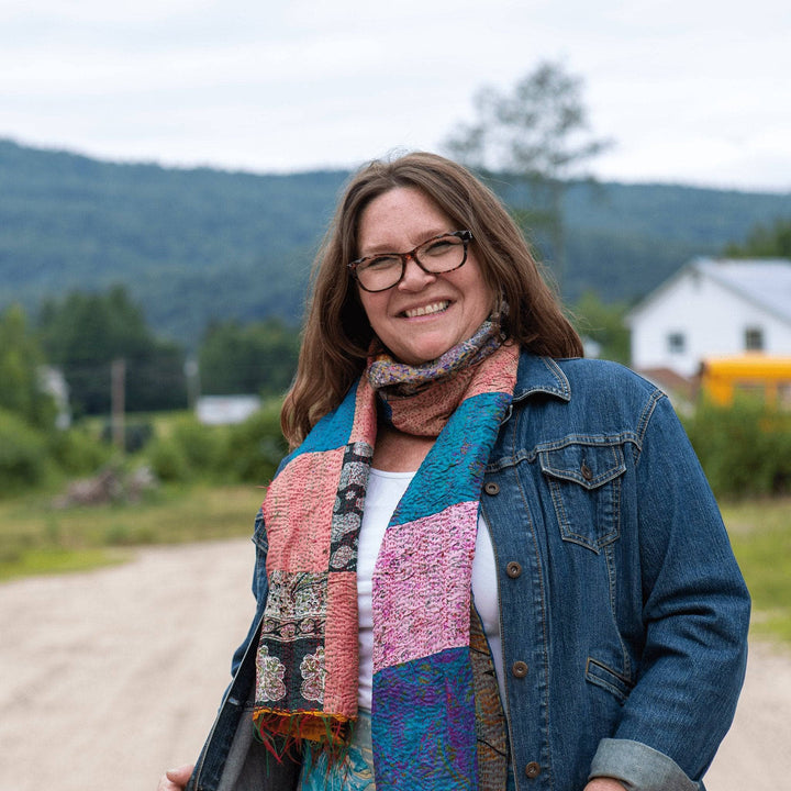 Smiling woman in a patchwork scarf and denim jacket, standing outdoors in a rural area.