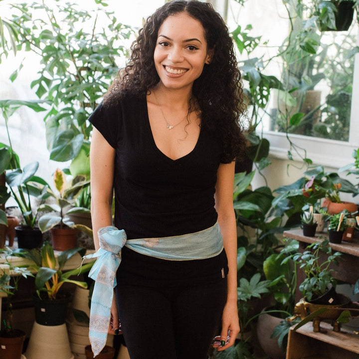 Model wearing light blue boho vintage sash accessory as a belt with a bow tied on one side with greenery in the background.