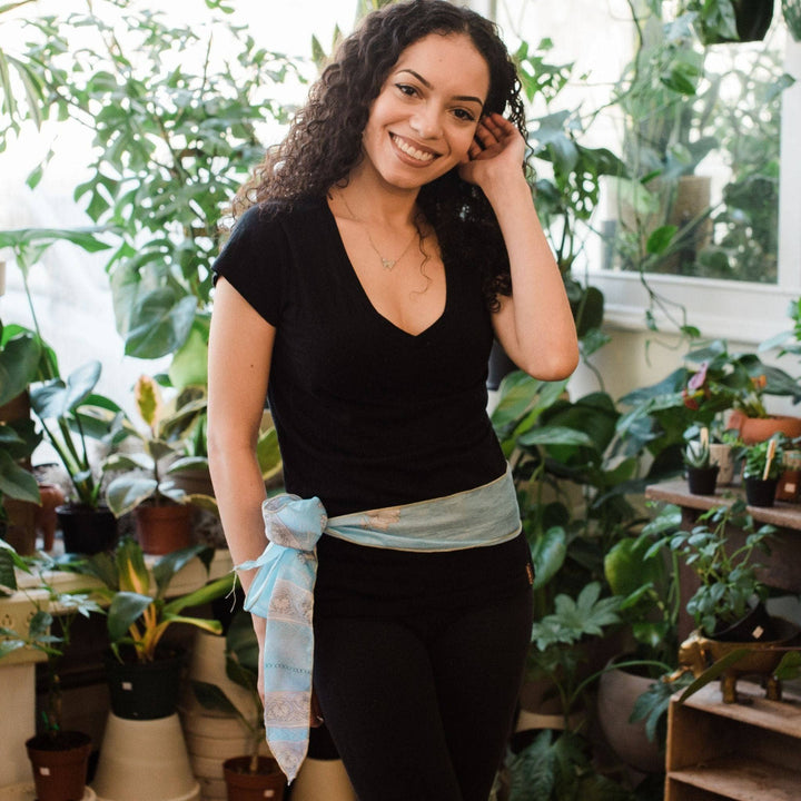 Model wearing light blue boho vintage sash accessory as a belt with a bow tied on one side with greenery in the background.