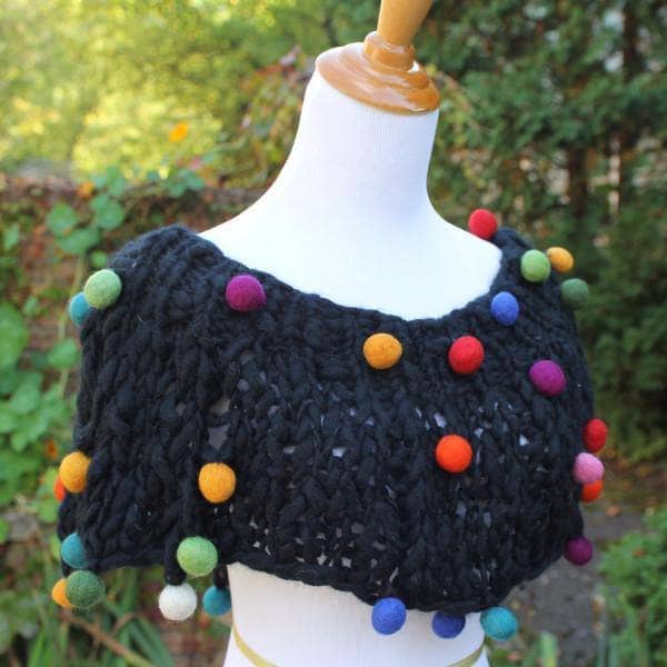 Mannequin wearing a Capowl Cowl in black with multicolored felt ball accents standing in front of greenery